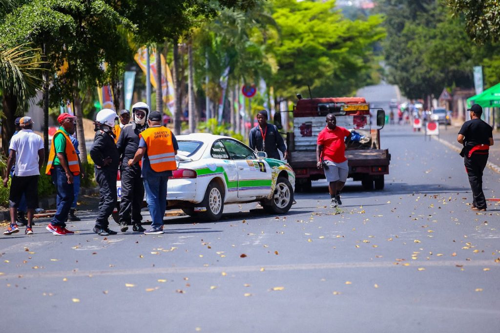 Rally event with crew and rally car on city street, featuring a tow truck and vibrant urban scene.